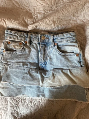Ljusblå jeanskjol med rå kant - Säljer en ljusblå jeanskjol med klassisk femficksdesign och rå, fransig kant nertill. Kjolen har bälteshällor, knapp och dragkedja framtill samt bakfickor. Perfekt nu till sommaren!