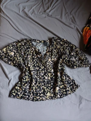 Svart blommig blus från Lindex - Blus med svart botten och småblommigt mönster i beige, vitt och ljusgult. V-ringad med knappar framtill och lätt puffade korta ärmar. Avslappnad passform och mjukt, tunt tyg som känns skönt mot huden. Passar perfekt nu inför sommaren🌺