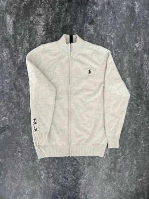 Ljusgrå ziptröja från Polo Ralph Lauren - Snygg ljusgrå stickad ziptröja från Polo Ralph Lauren med hög krage och ribbade muddar. Tröjan har RLX-brodyr på ärmen och den klassiska loggan på bröstet. Perfekt för lager på lager och enkel att matcha till olika outfits.