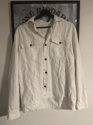 Vit overshirt med svarta knappar - Vit overshirt från Jack and Jones med svarta knappar och två bröstfickor med lock. Skjortan har klassisk krage och lång ärm, perfekt för lager-på-lager. Tillverkad i mjuk bomull för en avslappnad vibe. Storleken passar mer M än L.                             Skriv vid minsta fundering🍾