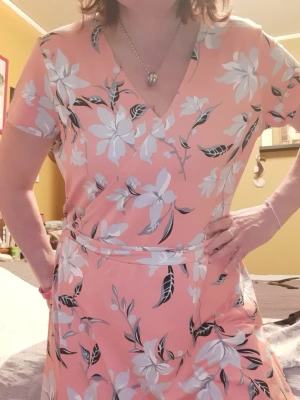 Persikofärgad blommig omlottklänning - Söt persikofärgad klänning med vita och svarta blommor. Klänningen har omlottmodell med v-ringning och korta ärmar. Midjebandet markerar figuren och tyget känns mjukt och följsamt. Perfekt för dig som gillar en feminin och fräsch stil. Storlek XXL 