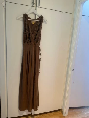 Ny beige Brun maxiklänning från H&M balklänning strl xs - Säljer en brun maxiklänning från H&M med djup v-ringning och dekorativa pärlor på axelpartiet. Klänningen har ett omlottliv och knytband i midjan. Materialet är mjukt och faller snyggt, perfekt för dig som gillar en bohemisk och chic stil. Helt ny Prislappen finns dock ej 
