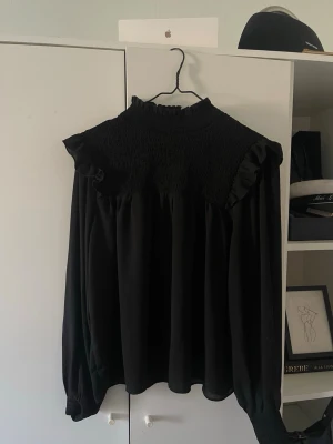 Svart blus med rynkad topp från Bik Bok - Säljer en svart blus från Bik Bok i storlek S. Blusen har rynkad topp med volangdetaljer vid axlarna och hög krage. Långa ärmar med lätt puff och smalare muddar. Materialet är lätt, mesh/lite genomskinlig och flowy.