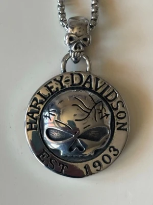 Harley-Davidson dödskalle halsband - Snyggt halsband i silverfärgad metall från Harley-Davidson med rund berlock prydd av en dödskalle och texten 'HARLEY-DAVIDSON EST 1903'. Kedjan har också en liten dödskalle vid fästet. Perfekt för dig som gillar tuffa accessoarer och bikerstil. Cirka 4*4cm och cirka 50gr 