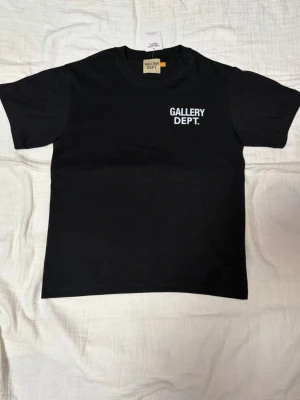 Svart t-shirt från Gallery Dept. - Svart t-shirt från Gallery Dept. med vit logga tryckt på bröstet. Klassisk rund halsringning och korta ärmar. Tillverkad i mjuk bomull för en skön och avslappnad stil. Perfekt för dig som gillar streetwear och enkel design.