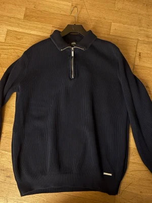 Mörkblå ribbad tröja med halv dragkedja - Snygg mörkblå ribbad tröja från ICON med halv dragkedja framtill och hög krage. Tröjan har lång ärm och en liten metallbricka vid nederkanten. Perfekt för dig som gillar stilrena och klassiska plagg.