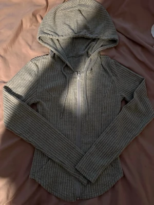 Beige ribbat set med hoodie och byxor - Mysigt loungewear-set i grå ribbad trikå. Setet består av en zip-hoodie med dragsko i huvan och långa ärmar samt matchande byxor med utsvängda ben. Perfekt för chill dagar hemma. Materialet är mjukt och stretchigt för extra komfort.