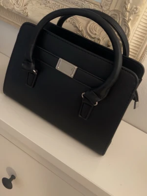 Svart handväska med silverdetaljer - Stilren svart handväska i slät konstläder med två rejäla handtag och silverfärgad metallplatta framtill. Väskan har ett rymligt huvudfack med dragkedja och innerficka för småsaker. Perfekt för dig som gillar klassisk och enkel design.