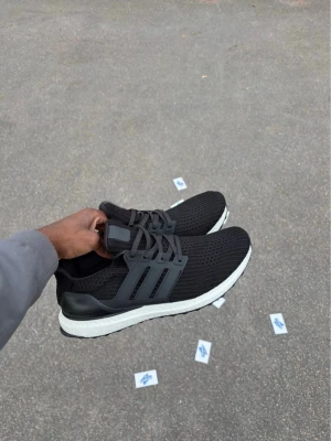 Adidas Ultraboost svarta sneakers - Säljer ett par svarta Adidas Ultraboost sneakers med mesh-överdel och grå detaljer på sidorna. Skorna har en vit Boost-sula som ger grym komfort och en sportig look. Perfekta för dig som gillar stilrena och bekväma sneakers.