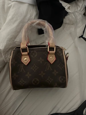 Monogram handväska - Säljer en snygg brun handväska med klassiskt monogrammönster och ljusa detaljer. Väskan har två stadiga handtag, guldfärgade metalldetaljer och dragkedja. Perfekt för dig som gillar lyxiga accessoarer och tidlös design.