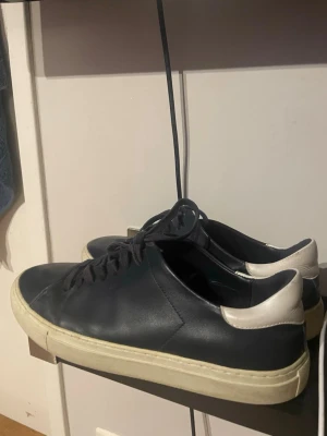 Svarta sneakers med vit häl - Stilrena svarta sneakers i läder med vit häl och vit sula. Klassisk låg modell med svarta skosnören och enkel design som passar till det mesta. Perfekta för dig som gillar minimalistisk stil.