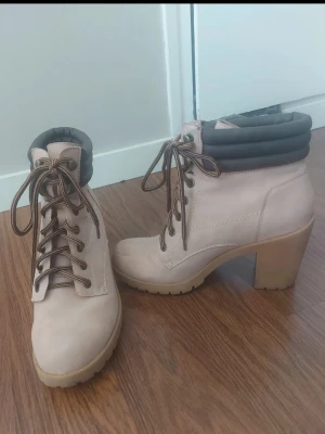 Beige stövletter med klack och snörning - Snygga beige stövletter med grov klack och snörning framtill. Skorna har en vadderad, mörkbrun kant upptill och robust sula med bra grepp. Perfekta för dig som vill ha både stil och höjd. Materialet ser ut att vara mocka eller liknande.