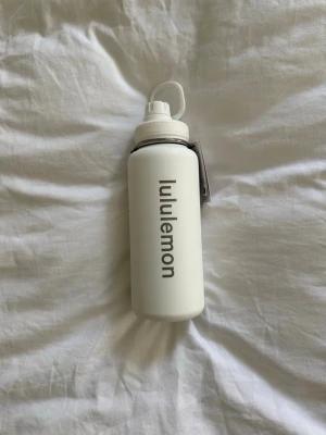 Vit vattenflaska från Lululemon - Snygg vit vattenflaska från Lululemon med svart logga längs sidan. Flaskan har ett praktiskt skruvlock med bärhandtag och är tillverkad i tåligt material, perfekt för träning eller gymmet. Denna är så snygg och helt oanvänd då jag har två stycken💓