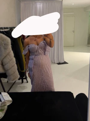 Ljuslila långklänning med paljetter - Säljer en elegant ljuslila långklänning med glittriga paljetter och offshoulder ärmar. Klänningen har en figurnära passform, markerad midja och vacker v-ringning. Perfekt för bal eller festliga tillfällen. Materialet känns lyxigt och skimrande.