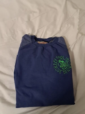 Petrolind - Snygg blå tröja med tryck i grönt på bröstet där det står 'Petrol Ind. Blue Denim' och ett coolt lejonmotiv. Tröjan har rund hals och är perfekt för en avslappnad stil.priset är 125 kroner