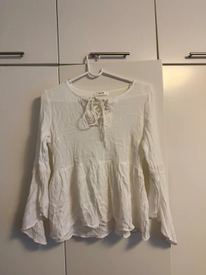 Vit boho-blus från batik - Söt sommar blus från Kaffe i boho-stil med långa ärmar och dekorativ knytning vid halsen. Blusen har volang nedtill och spetsdetaljer på ärmarna. Tillverkad i ett tunt, luftigt material som känns skönt mot huden. 