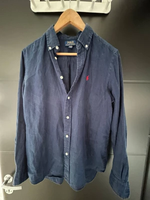 Marinblå linneskjorta från Polo Ralph Lauren - Klassisk marinblå skjorta i linne från Polo Ralph Lauren med knappar framtill och röd broderad logga på bröstet. Skjortan har lång ärm och button-down krage, perfekt för en stilren look.