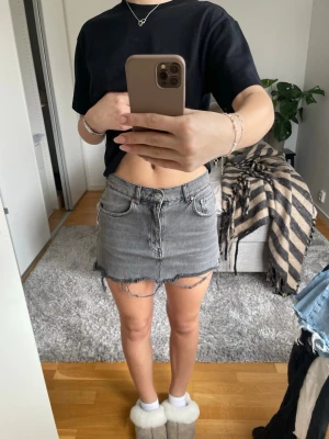 Grå jeanskjol med rå kant - Trendig grå jeanskjol med rå, fransad nederkant och klassiska fickor fram och bak. Kjolen har en kort passform och stängs med dragkedja och knapp framtill. Perfekt för dig som gillar en avslappnad och edgy stil. Den är lite sönder uppe vid kanten. Kolla bild 