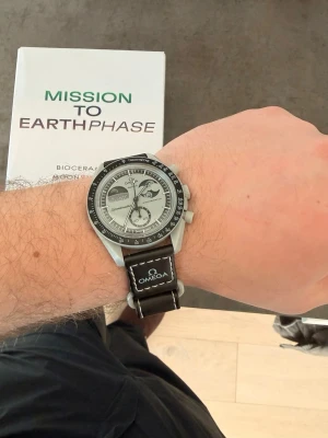 Omega x Swatch  - Snygg Omega x Swatch Speedmaster MoonSwatch Mission to Earthphase med silverfärgad urtavla och svart bezel. Klockan har svart band med vit söm och tydlig Speedmaster-logga. Biokeramisk boett och klassisk kronografdesign – perfekt för dig som gillar stilrena accessoarer.