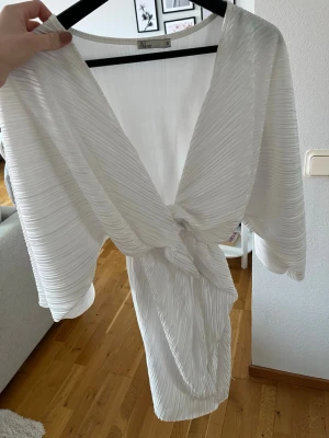 Vit plisserad omlottklänning från Nelly - Snygg vit plisserad klänning från Nelly med omlottmodell och knytning i midjan. Klänningen har vida  ärmar och ett skönt tyg som inte är genomskinligt. 