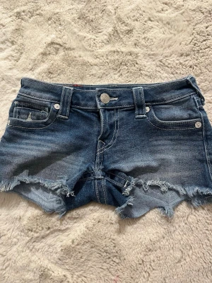 True Religion jeansshorts  - Säljer ett par blå jeansshorts från True Religion med rå franskant och slitningar. Shortsen har klassiska fem fickor, silverfärgade knappar och en patch med logotyp på baksidan. Perfekt för sommaren!