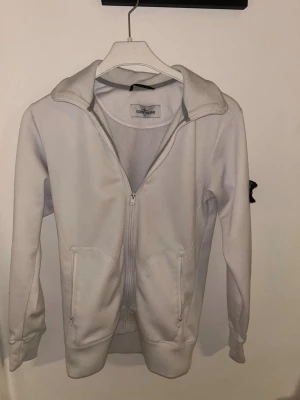 Vit zip hoodie från Stone Island - Säljer en clean vit hoodie från Stone Island med dragkedja framtill och två stora fickor. Klassisk logopatch på ärmen och ribbade muddar. Tillverkad i mjukt bomullsmaterial för skön känsla. Perfekt för dig som  vill ha något stilrent.
