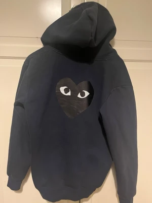 Mörkblå hoodie från Comme des Garçons - Säljer en mörkblå hoodie från Comme des Garçons PLAY med dragkedja, huva och fickor framtill. Ikoniskt hjärta med ögon på bröstet och stort tryck på ryggen. Skön bomullskvalitet och avslappnad passform. Perfekt för dig som gillar streetwear och snygga detaljer.