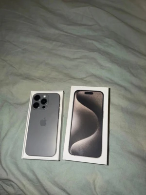 iPhone 15 Pro  titan grå - Säljer en iPhone 15 Pro i snygg titan grå färg. Telefonen har en stilren design med rundade kanter, tre kameror på baksidan och Apples logga. Materialet är en mix av glas och titan för en exklusiv känsla. Levereras i originalkartong. Mobilen är på 87% batteri hälsa.