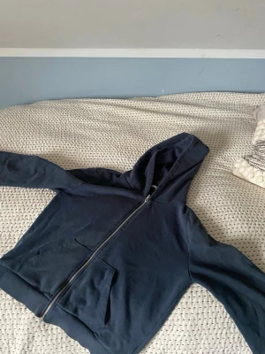 Mörkblå zip-hoodie med huva - Säljer en mörkblå hoodie med dragkedja och huva. Tröjan har två stora fickor framtill och ribbade muddar vid ärmslut och nederkant. Perfekt för dig som gillar en enkel och stilren look. Materialet är mjukt och bekvämt, passar till chill dagar. Passar till 11-13 år. 