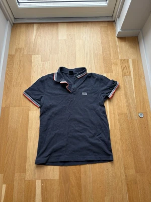 Mörkblå piké från Hugo Boss - Snygg mörkblå pikétröja från Hugo Boss med vit och orange rand på krage och ärmslut. Klassisk passform och diskret logga broderad på bröstet. Perfekt till jeans eller shorts för en clean look. Storlek S. Hör av dig vid frågor!