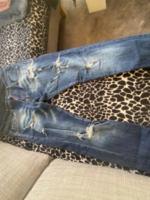 Blå slitna jeans med hål - Säljer ett par blå jeans med kraftiga slitningar och hål på flera ställen. Jeansen har en klassisk femficksmodell och är i en tvättad denim med ljusa partier. Perfekta för en avslappnad och trendig streetstil.