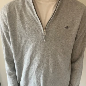 Grå GANT half zip tröja - Snygg grå långärmad tröja från GANT med half zip och broderad logga på bröstet. Tröjan har ribbad krage och muddar, tillverkad i mjuk bomull som är skön mot huden. Har ett litet hål i ena armhålan men är fixat. Säljs såklart för lägre pris pga det. Köpt på NK. Storlek 170 cm.