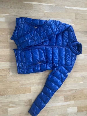 Blå dunjacka från Moncler - Snygg blå dunjacka från Moncler med glansigt yttertyg och quiltad design. Jackan har två lagningar som ni kan se på bild 2 och 3 men annars är den i bra skick. Den är liten i storleken  och passar M/L