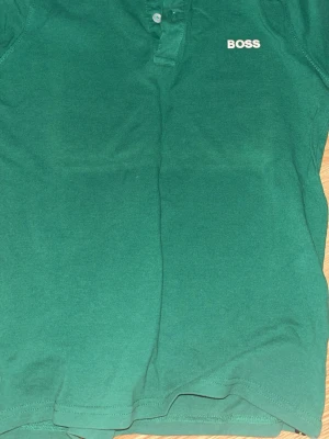 Grön pikétopp från BOSS - Storlek S /158 cm Snygg grön pikétopp med klassisk krage och knappar framtill. T-shirten har en enkel design. Perfekt till jeans eller shorts för en clean look.