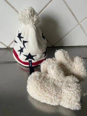 Vit mössa med stjärnor från Gant - Säljer en vit mössa från Gant med svarta stjärnor, röd rand och mörkblå knytband. Mössan har en stor tofs på toppen och är tillverkad i mjukt stickat material. Perfekt för dig som vill ha en snygg och bekväm accessoar till vintern. Barnmössa.  Illigs Vsntar med flercefoder