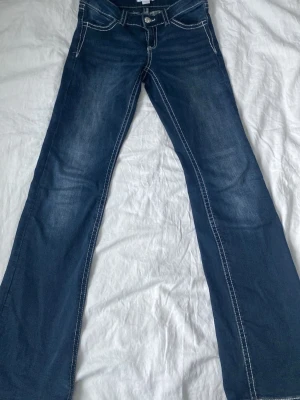 Blå bootcut jeans med kontrastsömmar - Snygga blå bootcut jeans med vita kontrastsömmar och klassisk femficksmodell. Jeansen har låg midja och är tillverkade i ett stretchigt denimtyg som ger en bekväm passform. Perfekta för en avslappnad och trendig look.