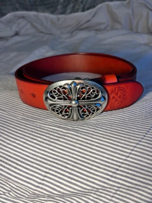 Chrome hearts bälte - Snyggt rött skärp från Chrome Hearts i skinn med ett stort, detaljerat silverfärgat spänne i korsdesign. Skärpet har en präglad logga vid änden och ger en cool statement-look till outfiten. 120 cm 