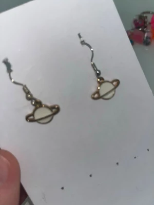 Guldiga örhängen med planetmotiv - Säljer ett par unika örhängen i form av planeter med ringar( handgjorda). De är guld- och vitfärgade och har en cool, minimalistisk design. Perfekta för dig som gillar rymdtema och vill sticka ut lite extra. Krokarna är i metall och själva planeten är vit med guldfärgad kant.