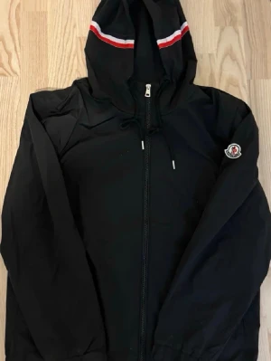 Svart vindjacka från Moncler - Svart vindjacka från Moncler med huva och dragkedja framtill. Jackan har vita och röda ränder på huvan samt Moncler-logga på ärmen. Tillverkad i lätt polyester, perfekt för blåsiga dagar. Snygg och sportig design med normal passform.