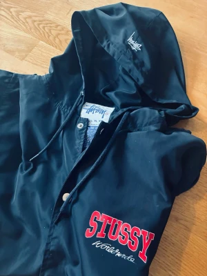 Svart vindjacka från Stussy med huva - Snygg svart vindjacka från Stussy med tryckt logga i rött och vitt på bröstet samt broderad logga på huvan. Jackan har knäppning framtill, dragsko i huvan och innerfoder med Stussy-mönster. Perfekt för streetwear-stil.
