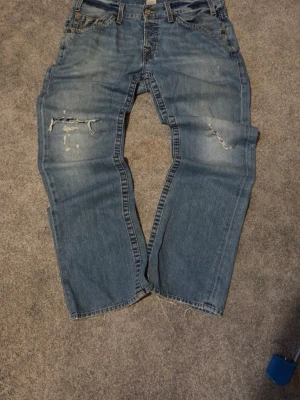 True religion jeans - Säljer ett par blå jeans med bootcut och slitningar på framsidan. Jeansen har klassisk femficksdesign och är tillverkade i denim. Perfekt för dig som gillar en avslappnad och trendig look.