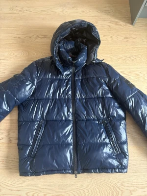 Michael kors jacka  - Snygg mörkblå pufferjacka med glansig finish och avtagbar huva. Jackan har två dragkedjeförsedda fickor fram och quiltad design. Perfekt för vinter, hösten och våren och ger en modern look. Dragkedja framtill och justerbara detaljer vid huvan. Pris går att diskuteras