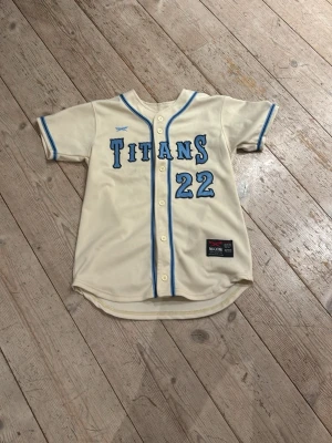 Beige basebolltröja Titans #22 - Snygg beige basebolltröja från Maxim med blå detaljer och texten 'Titans' samt siffran 22 på både fram- och baksidan. Knappar framtill och blå kantband längs ärmar och krage. Perfekt för sportiga tillfällen eller som streetwear.