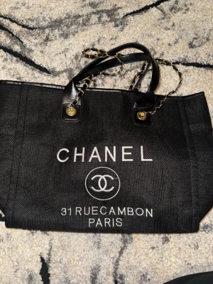 Svart Chanel handväska - EJ ÄKTA!! Snygg svart handväska från Chanel med vita logotypdetaljer och texten '31 Rue Cambon Paris' på framsidan. Väskan har dubbla handtag i läder med guldfärgade kedjor och insida med svart foder. 
