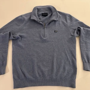 Blå halvzip-tröja från lexington - Snygg ljusblå stickad tröja från lexington med halv dragkedja och klassisk krage. Tröjan har ribbade muddar och en liten logotyp på bröstet. Perfekt för en avslappnad men stilren look.