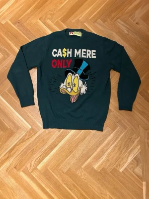 MC2 Saint Barth tröja - Mörkgrön stickad MC2 Saint Barth tröja med tryck av Joakim von Anka och texten 'CASH MERE ONLY' i vitt, svart, gult och rött. Tröjan har rund hals, långa ärmar och ribbade muddar. Perfekt för dig som gillar statement-plagg och tecknade motiv.