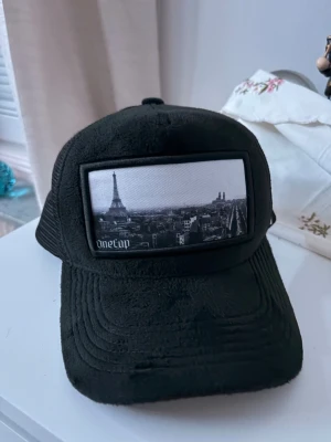 Svart truckerkeps med Paris-motiv - Svart truckerkeps från OneTap med mesh baktill och ett stort tygmärke framtill med svartvit bild på Eiffeltornet och Paris skyline. Kepsen har böjd skärm och justerbar passform. Perfekt för en cool streetstyle-look.