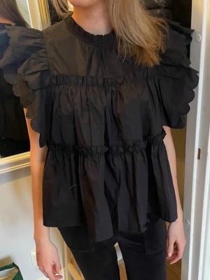 Svart volangblus med puffärm - Trendig svart blus med volanger och puffiga korta ärmar. Blusen har rund halsringning och är tillverkad i ett lätt, vävt material som ger en luftig känsla. 