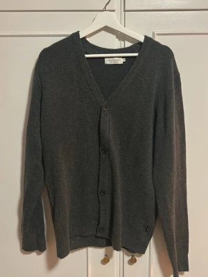 Mörkgrå kofta från Casual Friday - Mysig mörkgrå kofta från Casual Friday med v-ringning och knäppning framtill. Långärmad modell i mjukt stickat material, perfekt att slänga över en t-shirt eller skjorta. Klassisk och enkel design som passar till det mesta.