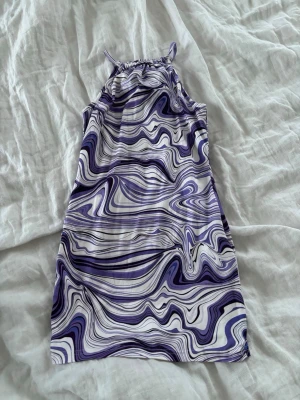 Lila och vit swirlmönstrad klänning - Säljer en kort klänning med swirlmönster i lila, vitt och svart. Klänningen har halterneck med knapp i nacken och är figurnära i modellen. Perfekt för dig som gillar färg och unika mönster. Den är stretchig💜
